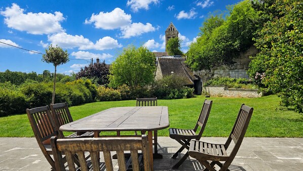 A patio with a wooden table and chairs, a stone wall, and a church tower in the background. - Country house, privacy and nature. (Lesges)