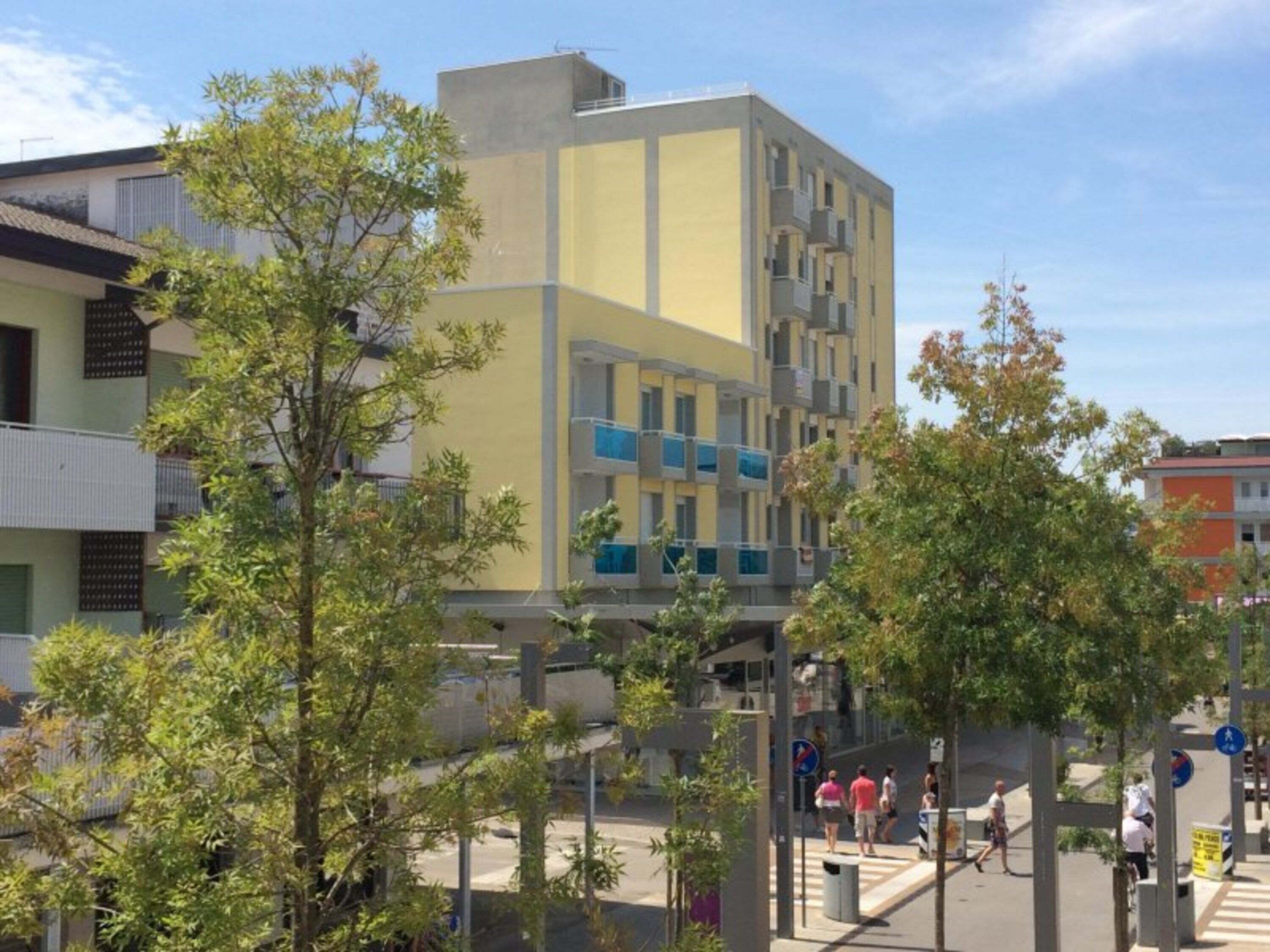 Un edificio moderno con fachada plana, varios balcones y una zona peatonal con árboles y un paso de cebra.