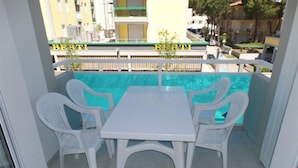 Un balcón con una mesa y sillas blancas, una piscina al fondo y un edificio con el letrero 'BERTI'.