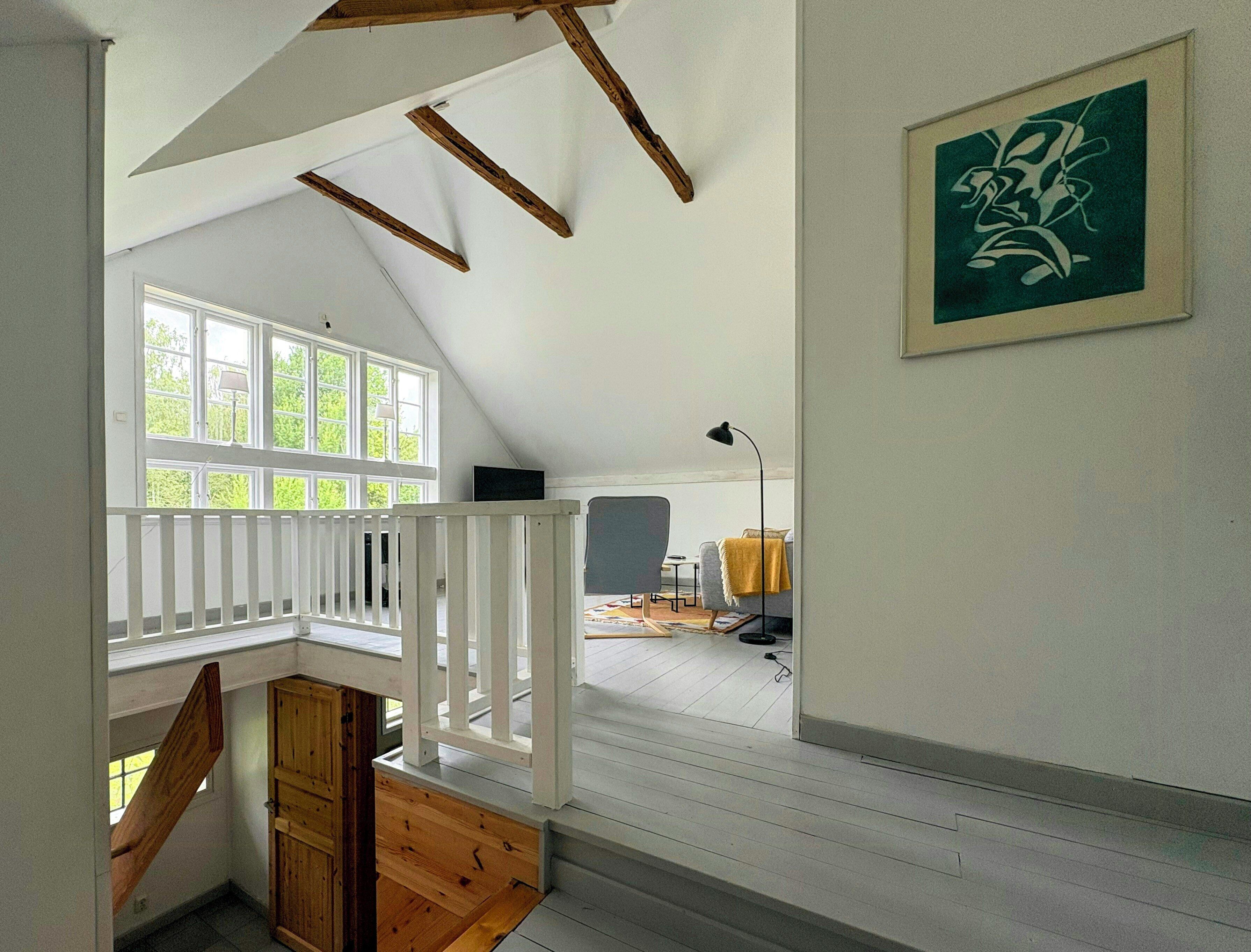 Um quarto com teto inclinado, vigas de madeira, um corrimão branco, uma janela e uma obra de arte emoldurada na parede.