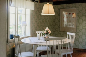 A dining area with a round table, white chairs, a vase with flowers, a hanging lamp, and a window with curtains. - 1909 farmhouse cottage (Vinslöv)