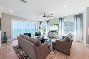 A spacious living room with a ceiling fan, two sofas, a coffee table, and a view of the ocean through large windows. - Beachfront Bliss by Beach Boutique Rentals (Anna Maria)