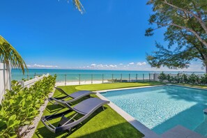 A swimming pool with lounge chairs, a view of the ocean, and a white fence. - Beachfront Bliss by Beach Boutique Rentals (Anna Maria)