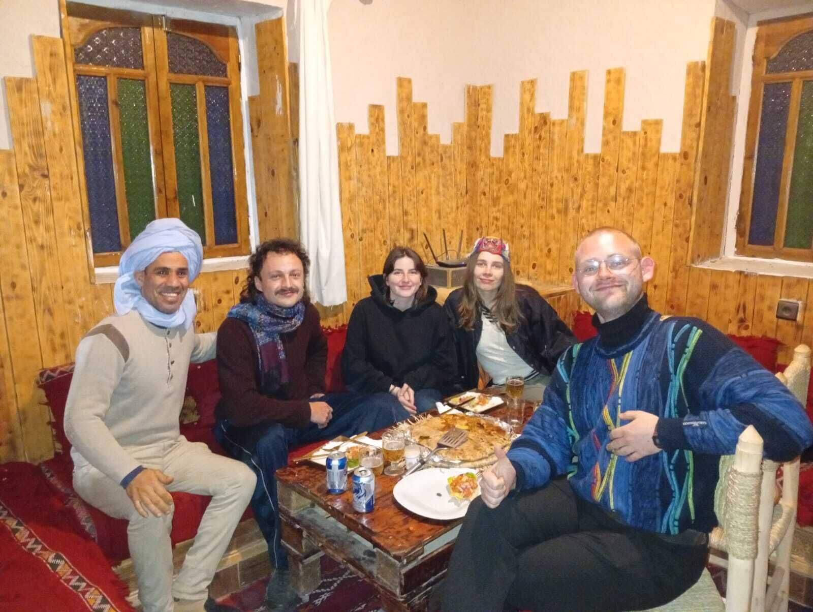 A group of people seated around a table with food and drinks, in a room with wooden walls and windows.