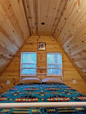 A wooden cabin room with a bed, two windows, and a ceiling light fixture.