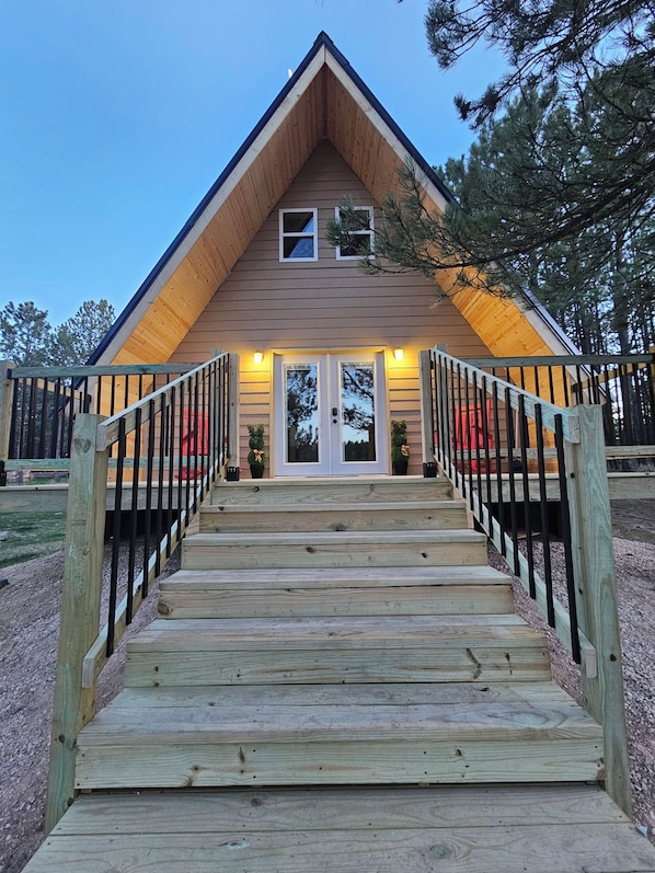 A wooden cabin with a steeply pitched roof, a front porch with a railing, and a set of stairs leading to the entrance.