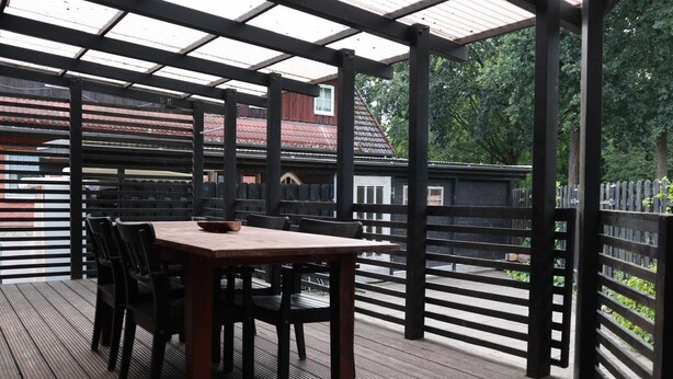 Un patio al aire libre con una mesa y sillas de madera, barandillas de metal y vista a un edificio con techo rojo.