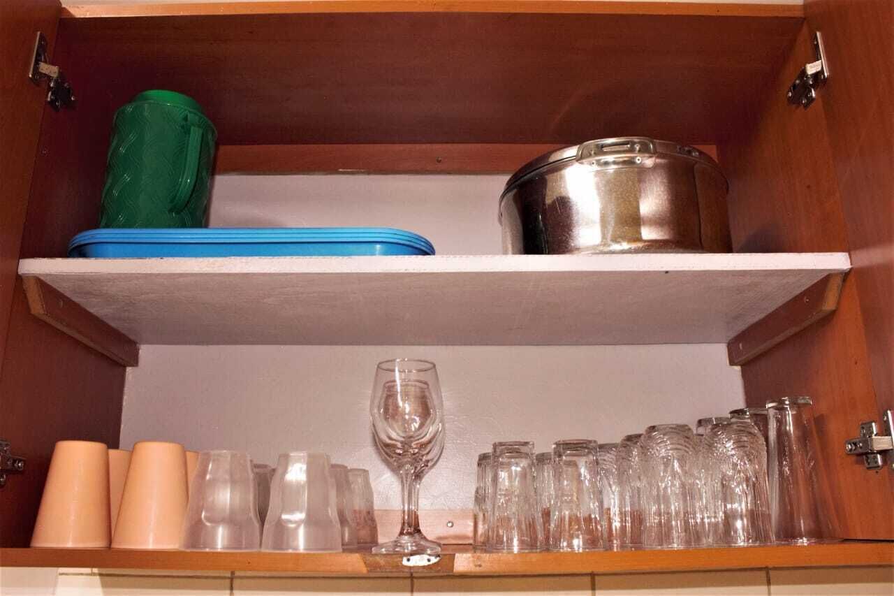 A kitchen cabinet with a shelf containing a green container, a blue container, a stainless steel pot, and various glassware including cups and a wine glass.