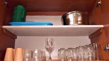 A kitchen cabinet with a shelf containing a green container, a blue container, a stainless steel pot, and various glassware including cups and a wine glass.