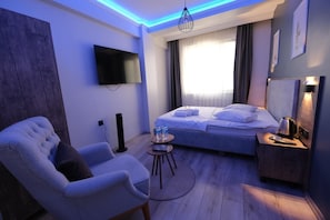 A hotel room with a bed, a chair, a small table, a TV, and a bedside table with a lamp. - Luna Suites Hotel (Eskişehir)