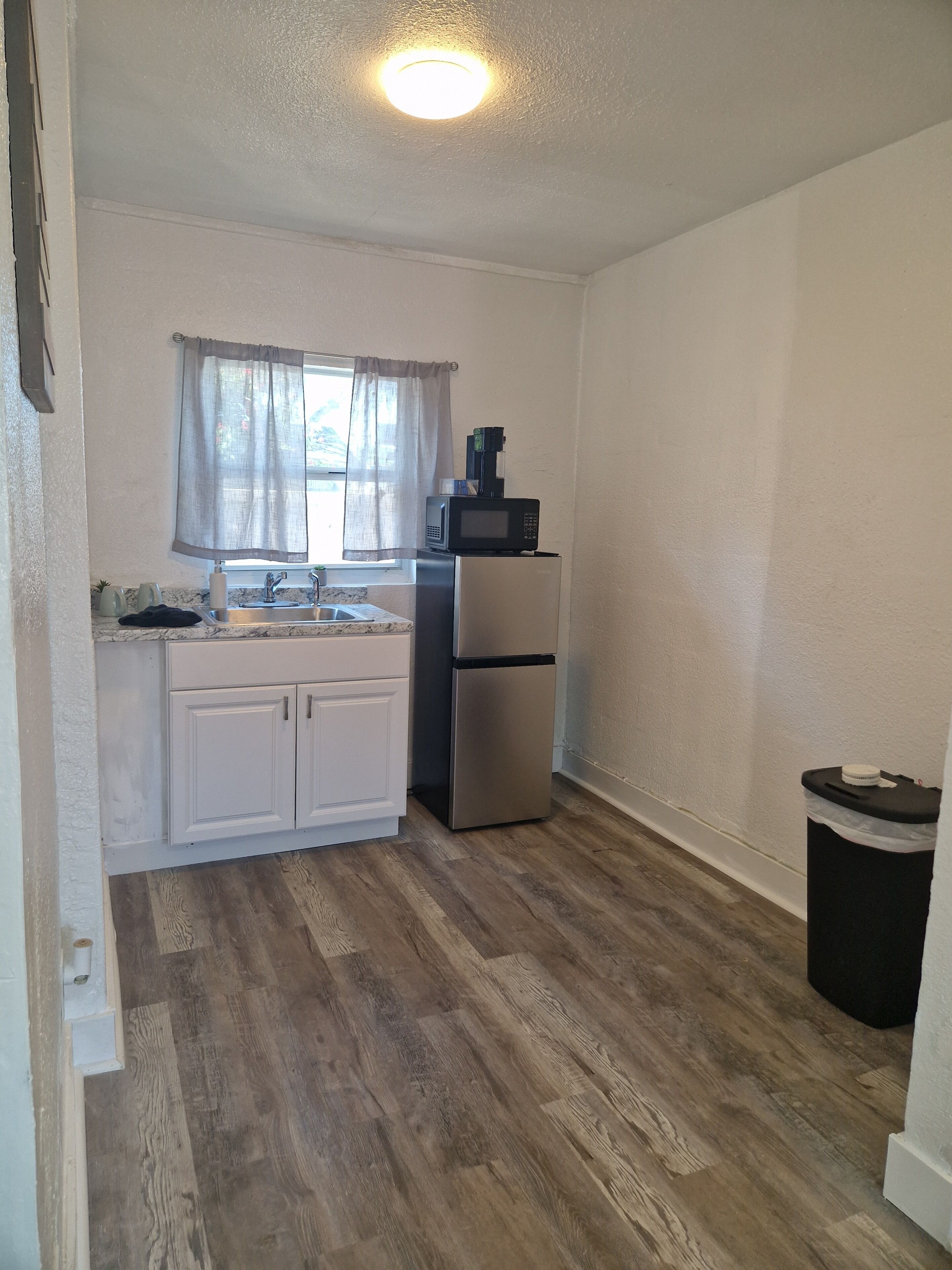 A compact kitchen with a refrigerator, microwave, sink, and window with curtains.