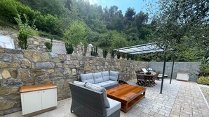 Une terrasse avec des murs en pierre, un canapé gris, une chaise en osier, une table basse en bois et une pergola.