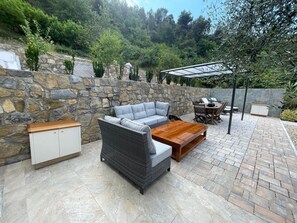 A patio with stone walls, a grey sofa, a wicker chair, a wooden coffee table, and a pergola. - Mimosetta Guest House: A Cozy Cottage Surrounded by Peace and Greenery, with Views of the Sea and Mountains (Vallebona)