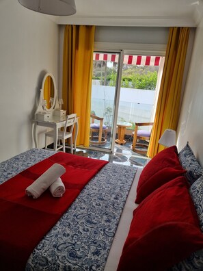 A hotel room with a bed, a desk with a mirror, a chair, and a view of a balcony with yellow curtains. - 1 bedroom seaside apartment (El Jadida)