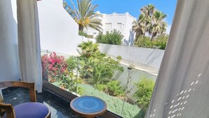 Ein Balkon mit zwei Holzstühlen, einem kleinen Tisch, einem gestreiften Sonnenschirm und Blick auf Gebäude und Grünflächen.