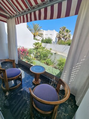 A balcony with two wooden chairs and a small table, a striped awning, and a view of buildings and greenery. - 1 bedroom seaside apartment (El Jadida)