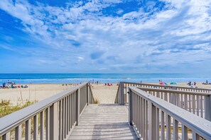 Ein hölzerner Steg führt zu einem Strand mit Menschen und Sonnenschirmen.