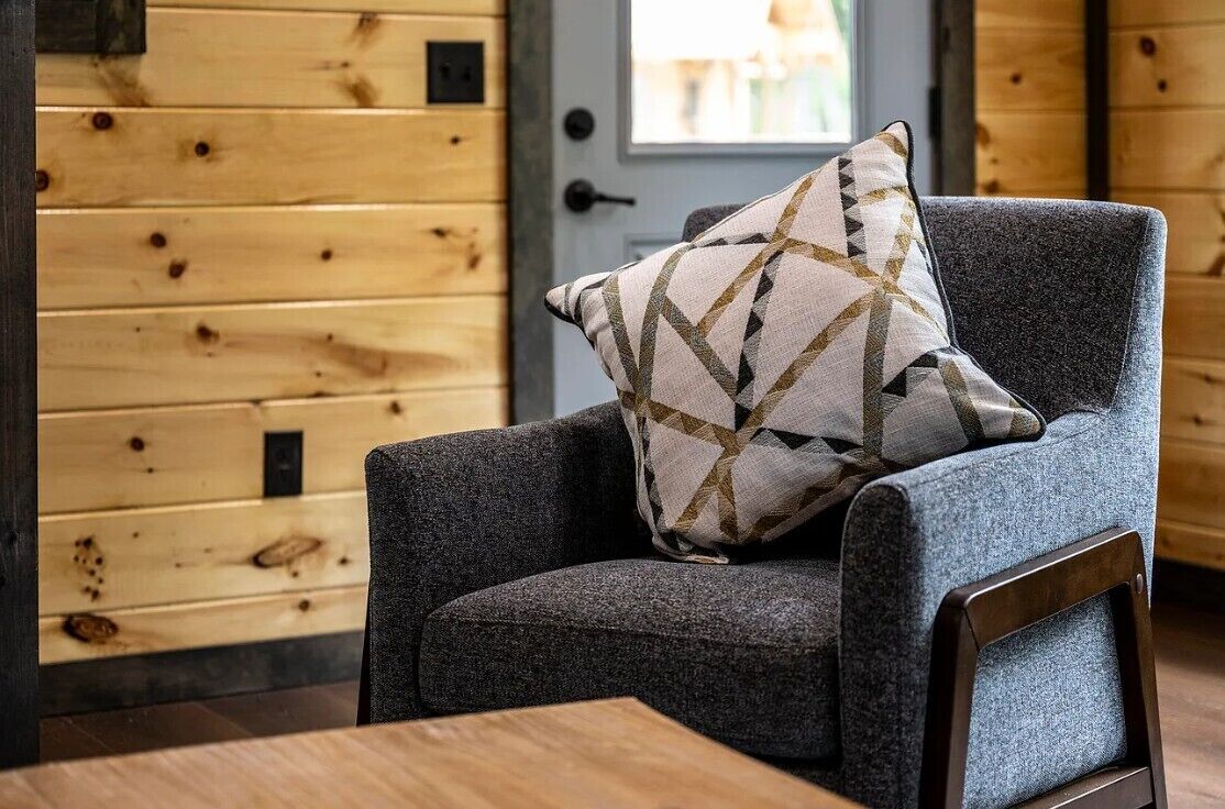 A grey armchair with a patterned pillow in a room with wooden walls and a door.
