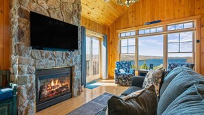 A stone fireplace with a TV mounted above it, a wooden ceiling, and a large window with a view of the outdoors. - Lodges 12 | Lake Views | Modern Condo (Rangeley)