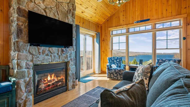 A stone fireplace with a TV mounted above it, a wooden ceiling, and a large window with a view of the outdoors. - Lodges 12 | Lake Views | Modern Condo (Rangeley)