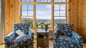 A room with two blue chairs with sailboat patterns, a small wooden table, and a window with a view of a house and mountains. - Lodges 12 | Lake Views | Modern Condo (Rangeley)