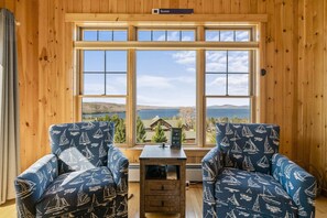 Ein Zimmer mit zwei blauen Stühlen mit Segelbootmuster, einem kleinen Holztisch und einem Fenster mit Blick auf ein Haus und Berge.