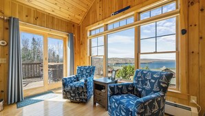 A wooden interior with large windows, two patterned armchairs, a small table, and a view of a lake. - Lodges 12 | Lake Views | Modern Condo (Rangeley)