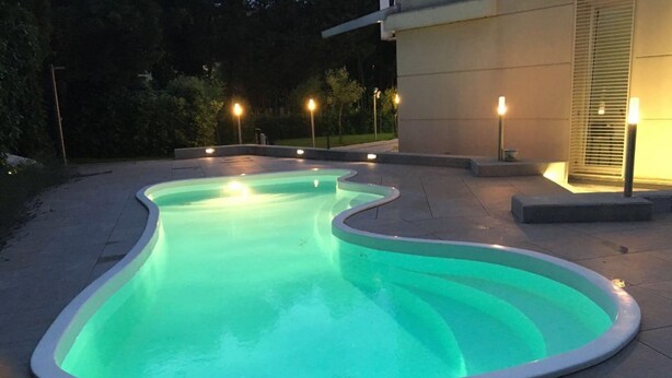 A swimming pool with a unique wavy edge design, surrounded by a tiled deck and outdoor lighting. - Villa Celeste Flexrent - Abissinia Real Estate (Scacciano)