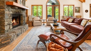 A cozy living room with a stone fireplace, leather furniture, and large windows. - 3BR, 3BA Mountain home with sweeping views in Waynesville. (Waynesville)
