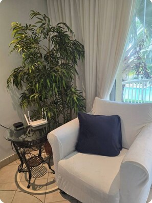 Un sillón blanco con un cojín oscuro, una mesa de vidrio con un router y una planta verde grande en una habitación con vistas al exterior.