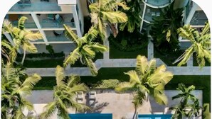 An aerial view of a resort with a central swimming pool, surrounded by palm trees and lounge areas.