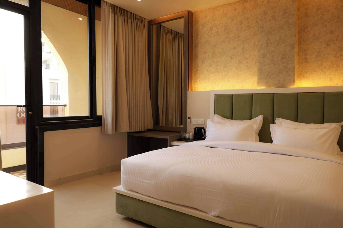 A hotel room with a large bed, a nightstand, a coffee maker, and a view through a window with curtains.