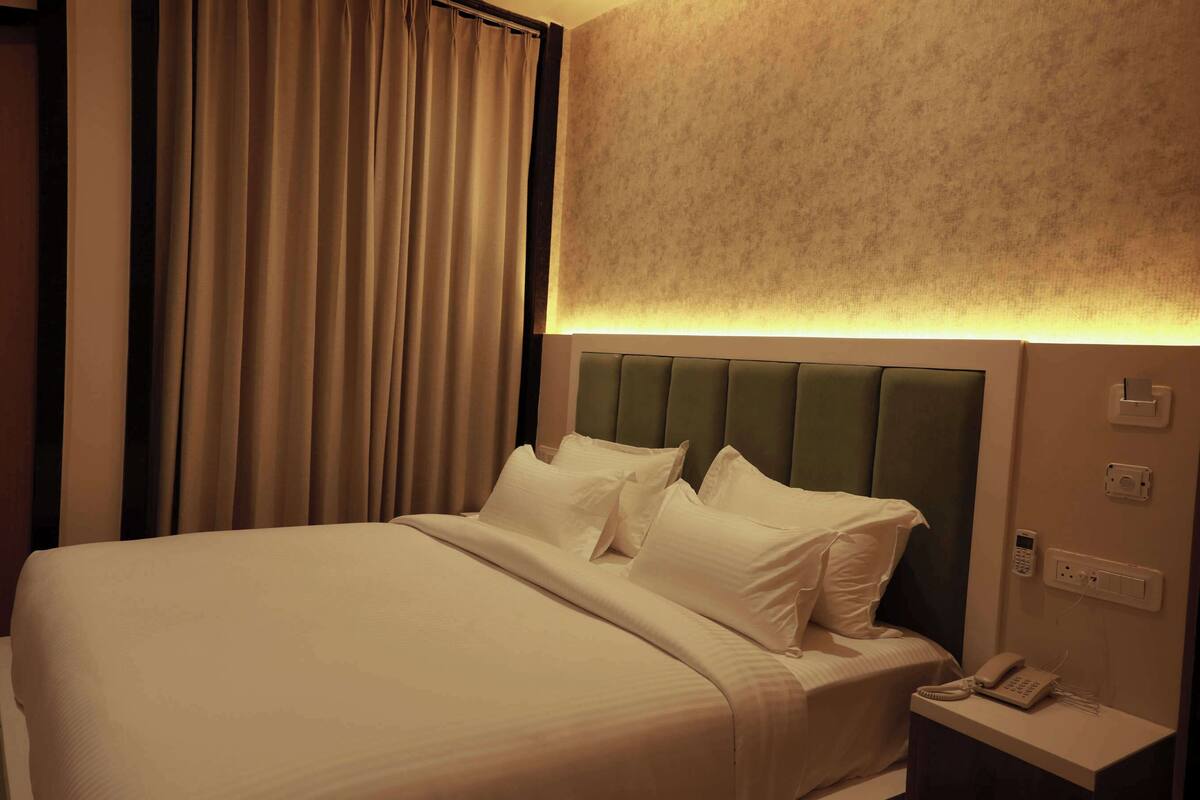 A hotel room with a bed, curtains, a headboard, a bedside table with a phone, and a wall-mounted light.