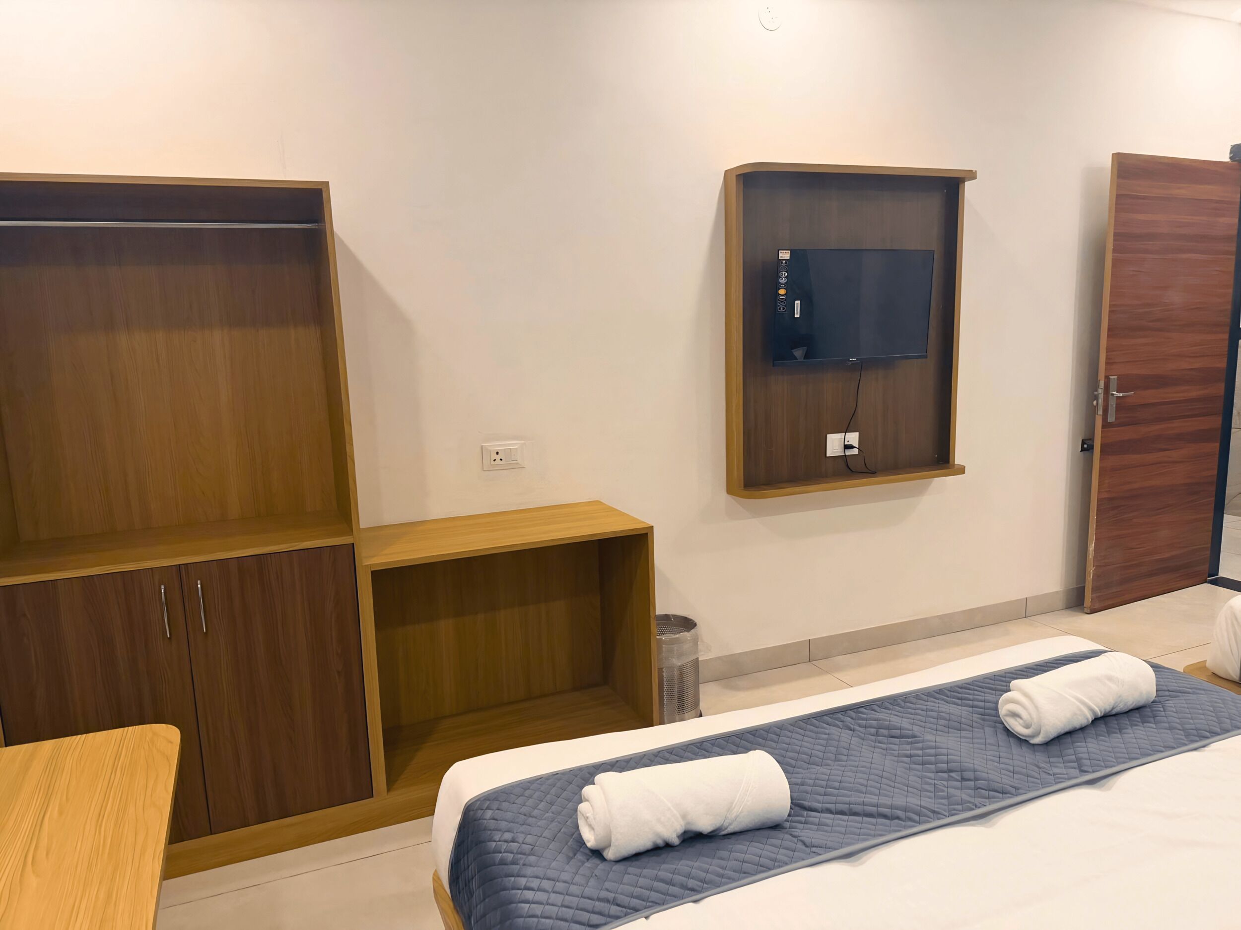 A hotel room with a bed, a wooden wardrobe, a television mounted on the wall, and a door.