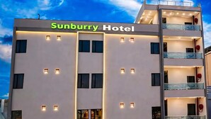 Un edificio moderno de hotel con un letrero que dice 'Sunburry Hotel'. Cuenta con varios pisos con balcones, una entrada de garaje y una fachada plana.