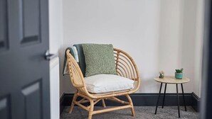 Una silla de mimbre con un cojín, una mesita redonda con una planta, y una puerta al fondo.