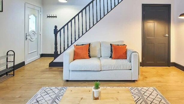 Una sala de estar con una escalera, un sofá con cojines, una mesa de centro con una planta y una puerta.