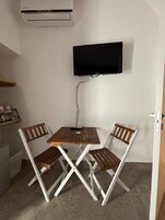 Una habitación pequeña con una mesa, dos sillas, un televisor de pantalla plana en la pared y un equipo de aire acondicionado sobre la mesa.