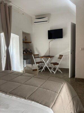 A bedroom with a bed, a TV mounted on the wall, a desk with chairs, and an air conditioning unit.
