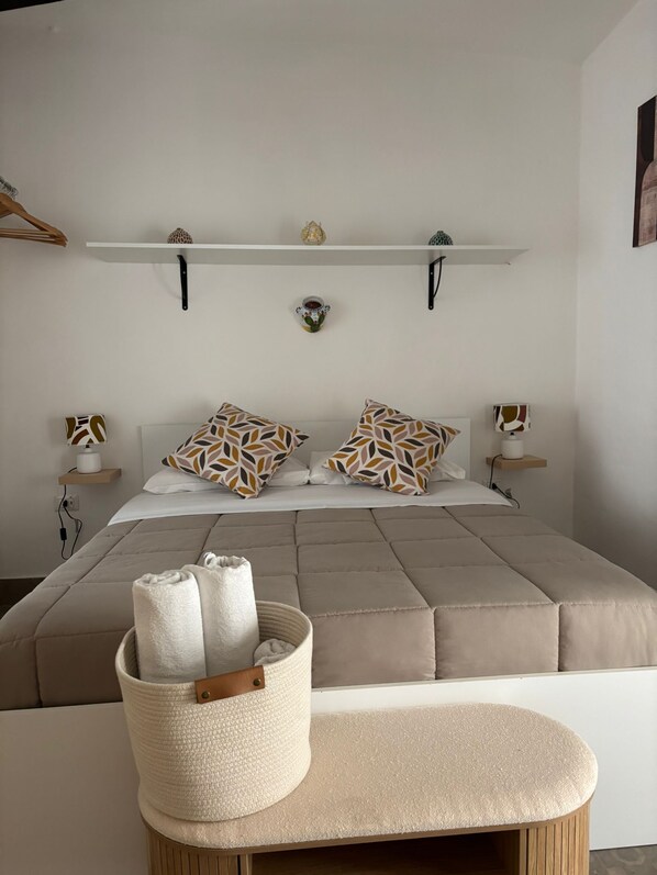 A bedroom with a bed, pillows, a bedside table, a lamp, a shelf with decorative items, and a basket with towels.