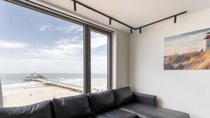 Una sala de estar moderna con un sofá de cuero negro, ventanas amplias con vistas a la playa y al muelle, y un cuadro en la pared.