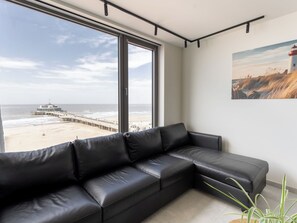 Ein modernes Wohnzimmer mit einer schwarzen Ledersofa, großen Fenstern mit Blick auf Strand und Steg, und einem gerahmten Bild an der Wand.