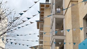 A multi-story residential building with balconies, a paved street, parked cars, and a row of informational posters on the right. - 10 Mins to Takism - Central & New Modern Flat (Istanbul)
