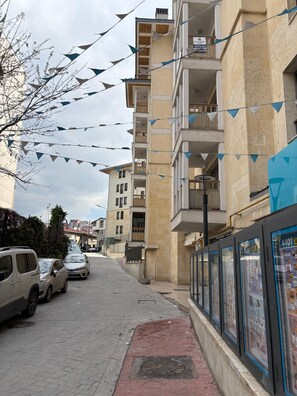 A multi-story residential building with balconies, a paved street, parked cars, and a row of informational posters on the right. - 10 Mins to Takism - Central & New Modern Flat (Istanbul)