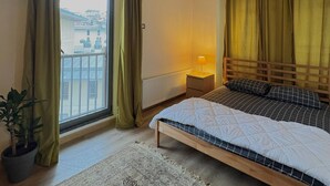 A bedroom with a bed, a nightstand, a lamp, a potted plant, a window with curtains, and a view of buildings outside. - 10 Mins to Takism - Central & New Modern Flat (Istanbul)
