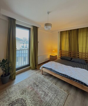 A bedroom with a bed, a nightstand, a lamp, a potted plant, a window with curtains, and a view of buildings outside. - 10 Mins to Takism - Central & New Modern Flat (Istanbul)
