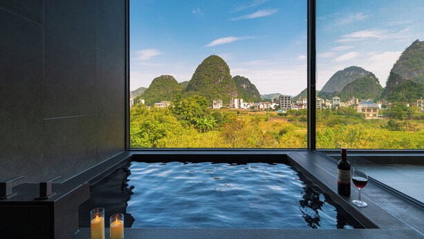 Un espacio de spa con un ventanal grande, una bañera de hidromasaje, velas y una botella de vino.