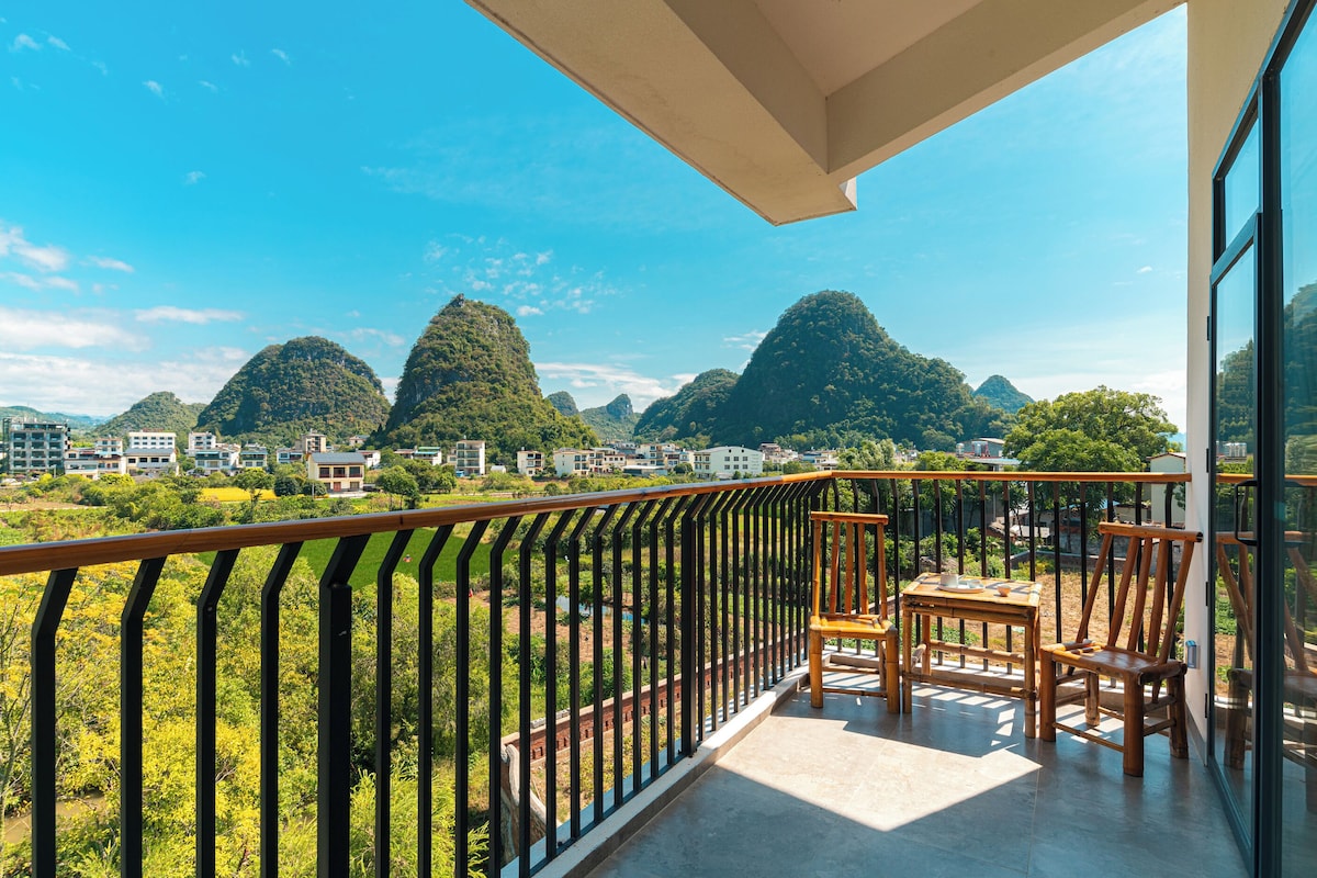 A balcony with a view of a mountainous landscape, a table with chairs, and a railing.