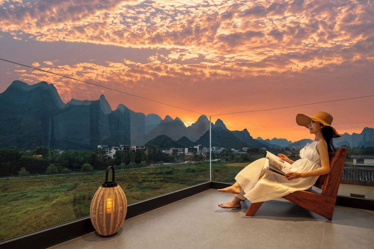 Una mujer sentada en un banco de madera con vistas a montañas y al atardecer.