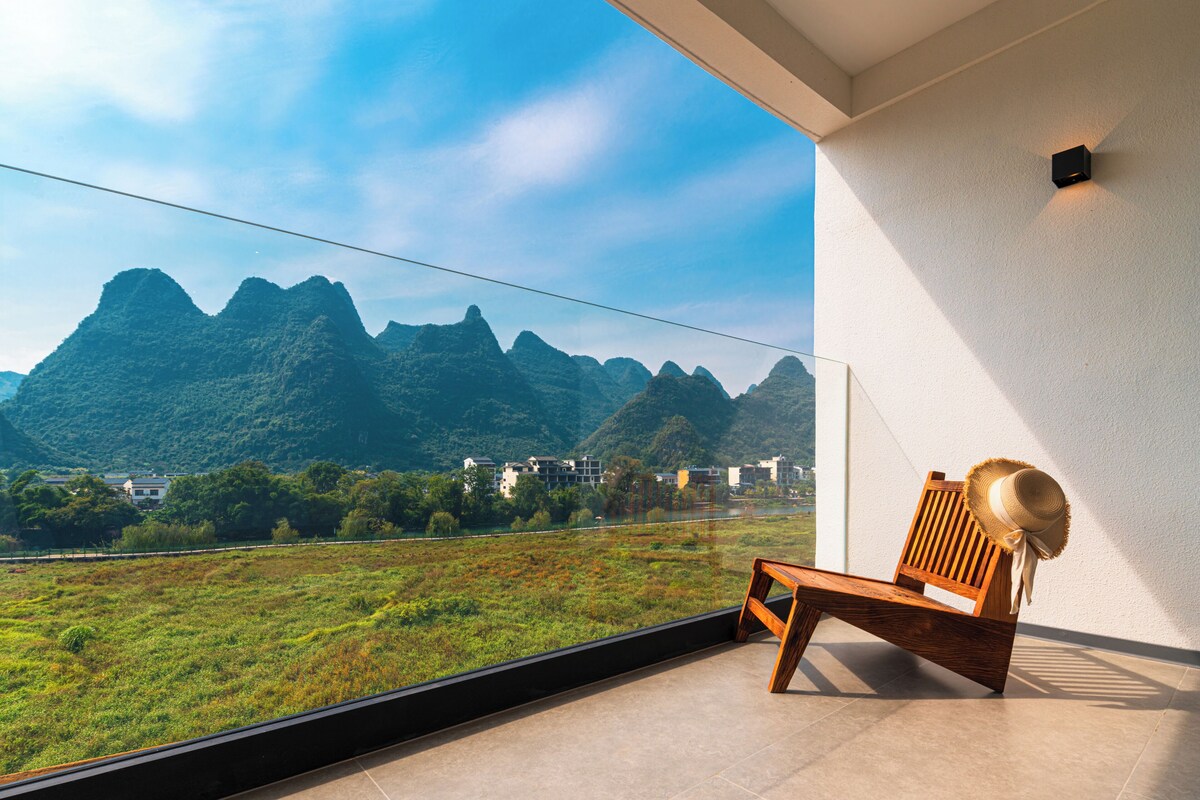 A balcony with a wooden chair and a straw hat, overlooking a mountainous landscape.